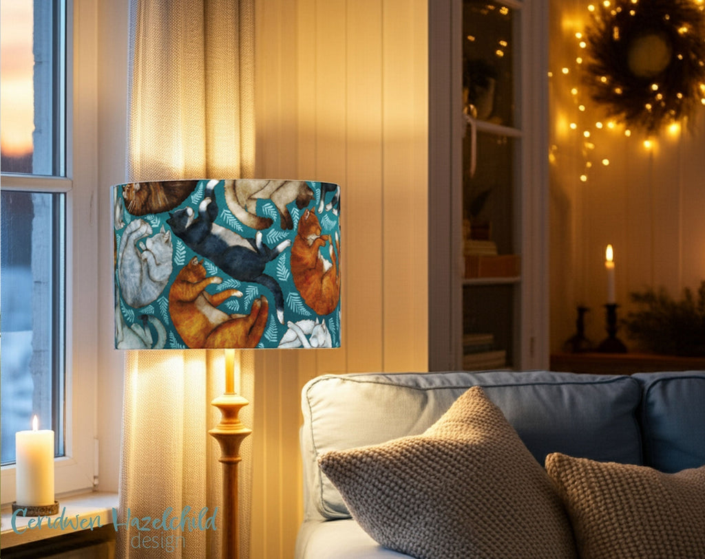 Illustrated drum lampshade featuring a pattern of sleeping cats in various colours on a teal background, glowing warmly in a softly lit living room with candles and fairy lights.
