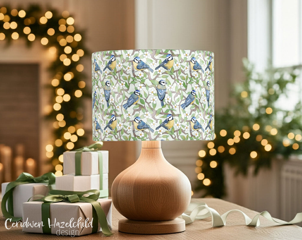 Organic cotton lampshade featuring hand-illustrated blue tits and blossom branches, displayed beside ribbon-tied gifts in a softly lit festive room. A whimsical woodland animal lampshade and ethical Christmas gift idea from a Cotswolds designer.