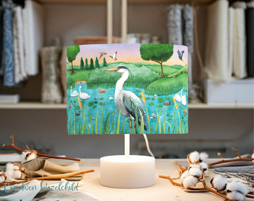 A lampshade with an image of a heron in a lake on it, standing on an artist studio surrounded by fabric and natural cotton plants.
