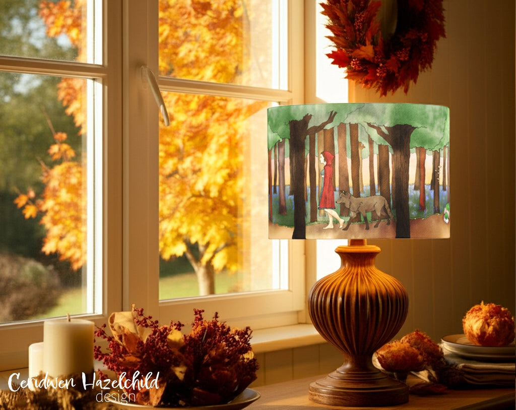 A lampshade with an illustration of Red Riding Hood and her wolf walking through the forest, on a table lamp decorated with autumnal decor.