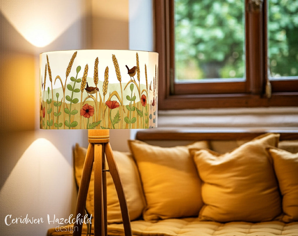 Organic cotton lampshade illustrated with wren birds, wheat stalks, and poppies, glowing warmly in a cosy reading nook with golden cushions. A handmade woodland animal lampshade designed for calm children’s bedrooms or nature-inspired home decor.