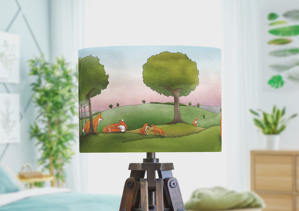 A woodland animal lampshade with foxes in the meadow full of trees under an evening sky. The fox lampshade rotates to show the wrap around scene, drawn by the artist.