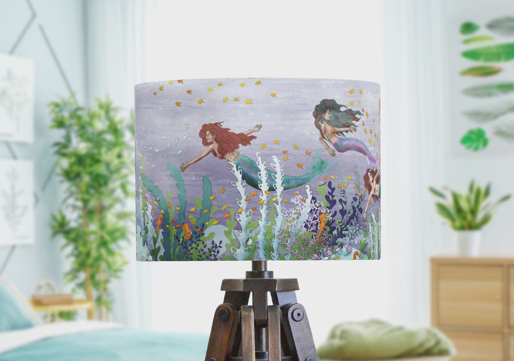 A mermaid lampshade showing an wrap around illustration of mermaids swimmign through a purple ocean and coral reef with orange fish, sea weed and seahorses. The handmade lamphade rotates to show the panoramic scene by the artist.