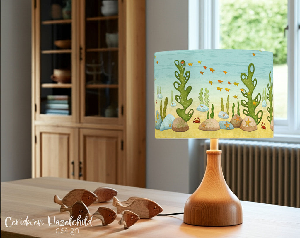 Under The Sea lampshade on an oak lamp base, styled on a wooden table with carved fish, in a bright, airy room with a bookcase and large window.
