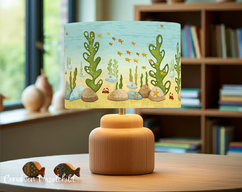 Under The Sea handmade lampshade in a light-filled office, placed on a wooden table with wooden fish decor, oak lamp base, and a bookcase in the background