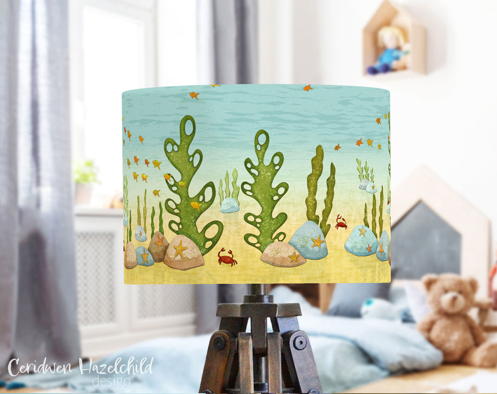 Under the Sea lampshade by Ceridwen Hazelchild on a tripod lamp base in a gender-neutral children’s bedroom, with a cosy bed, soft teddies, and a bright window in the background.
