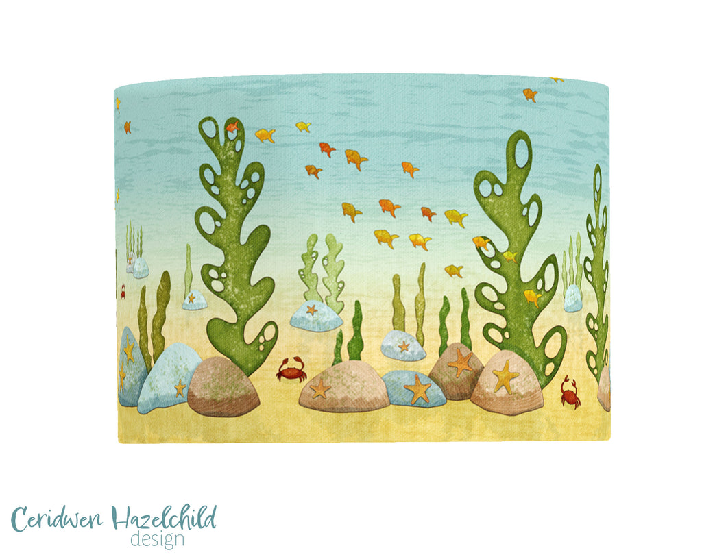Under the Sea lampshade by Ceridwen Hazelchild – handmade drum lampshade featuring an illustrated shallow ocean scene with fish, crabs, rocks, and seaweed in soft yellow and blue tones.