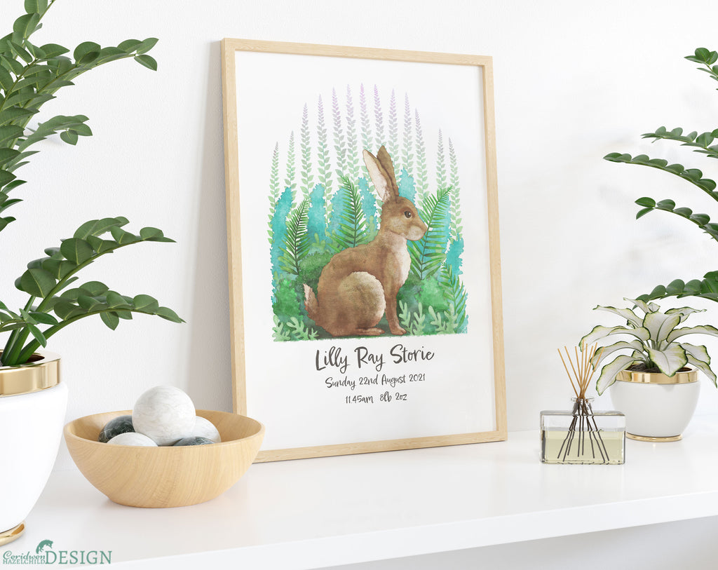 A framed illustration of a rabbit by Ceridwen Hazelchild, personalised with a baby girl's name and birth details, placed on a shelf and surrounded by plants.